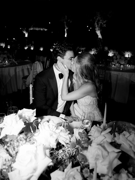 Bride and groom sharing a kiss while seated at reception table surrounded by white florals