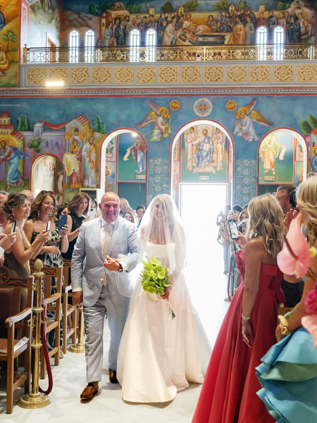 Bride entering colorful Orthodox church with her father during Athens wedding ceremony