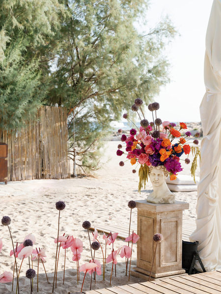 Bold pink and orange floral arrangement displayed on classical bust at beachfront wedding ceremony in Athens