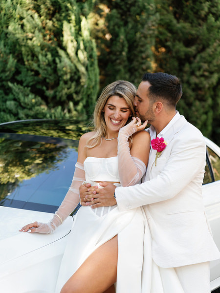 Bride and groom smiling beside white car following Greek Orthodox wedding ceremony