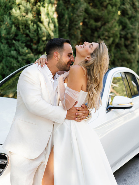 Groom in white suit embracing bride in couture gown beside wedding car in Athens