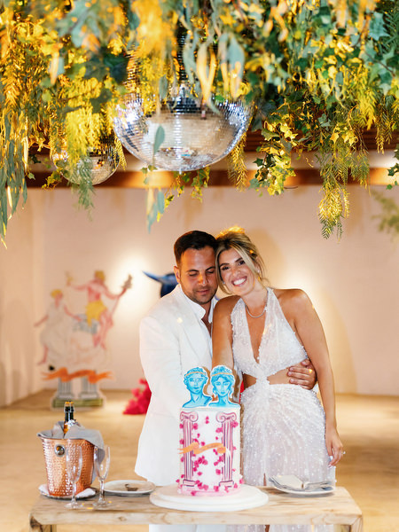 Bride and groom cutting colorful wedding cake beneath hanging greenery and mirrored balls in Athens