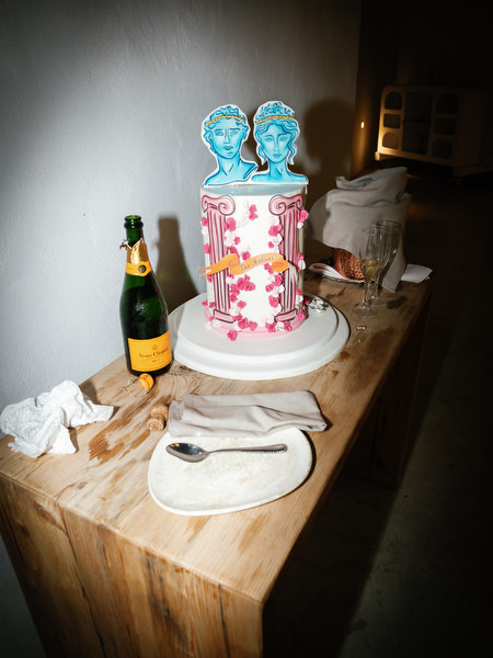 Modern wedding cake with graphic Greek-inspired topper beside champagne bottle on wooden reception table with editorial flash lighting