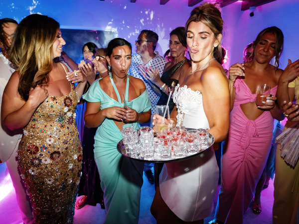 Bride carrying tray of champagne shots through packed wedding dance floor surrounded by bridesmaids in colorful dresses