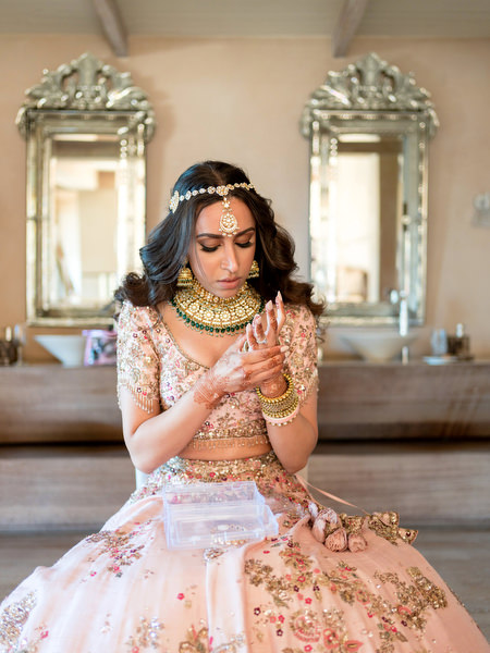 Bride adjusting gold jewellery while wearing powder pink embroidered lehenga at Private House Island Resort Athens Riviera Indian wedding.