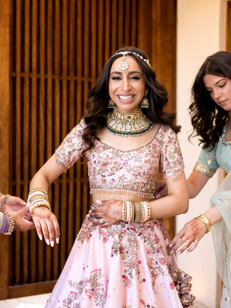Rebecca smiling in powder pink raw silk lehenga with intricate embroidery at colourful Indian wedding in Athens.