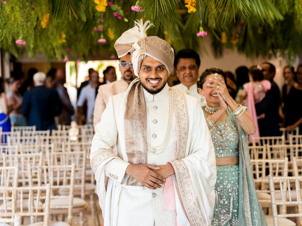 Groom smiling in ivory sherwani beneath floral mandap at Athens Riviera Indian wedding.