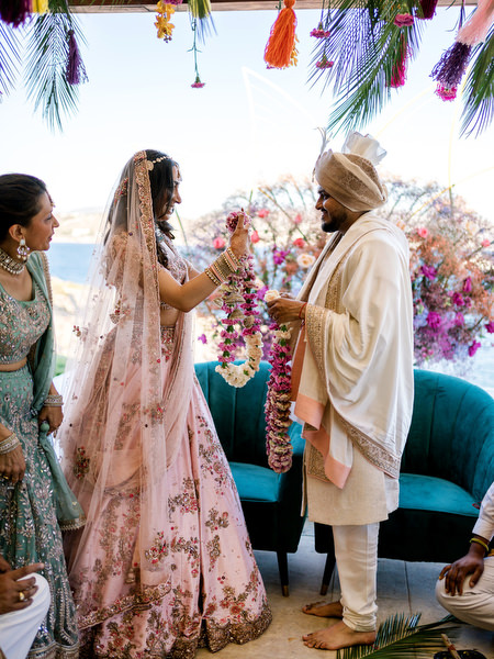 Bride and groom exchanging floral garlands under seaside mandap at colourful Indian wedding in Athens Riviera.