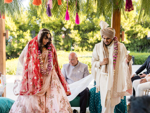 Bride and groom preparing for pheras under floral mandap at seaside Indian wedding in Athens.