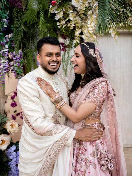 Bride and groom laughing together in powder pink lehenga and ivory sherwani at Athens Riviera wedding.