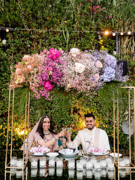 Bride and groom seated beneath pink and purple floral canopy at Athens Indian wedding reception.
