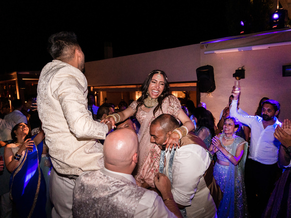 Bride and groom being lifted during energetic Bollywood dance floor moment in Greece.