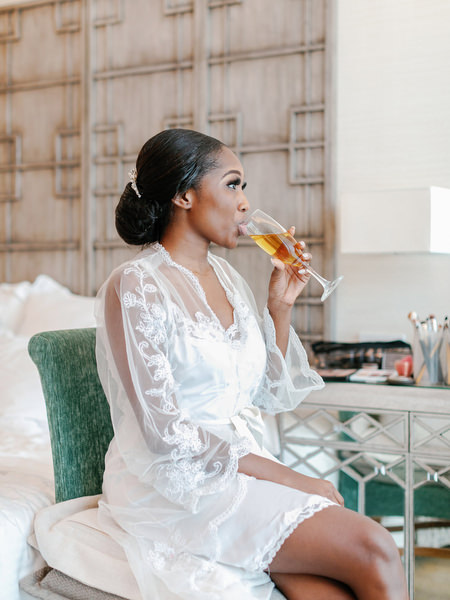 Bride in lace robe sipping prosecco during luxury Nigerian wedding morning at the Margi Hotel on the Athens Riviera.