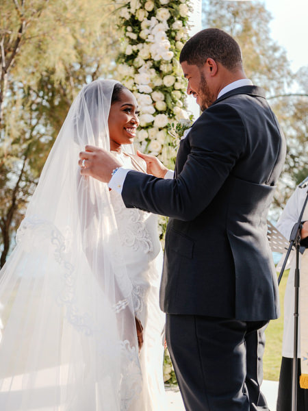 Groom lifting bride’s veil under floral arch during high-end Nigerian destination wedding ceremony at Ble Azure Athens Greece.