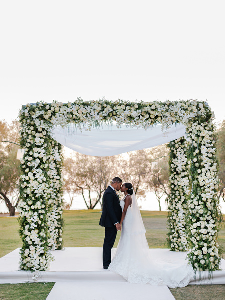 Bride and groom standing beneath white floral ceremony arch at Ble Azure Nigerian destination wedding in Greece.