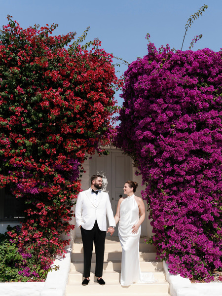 Bride and groom standing beneath blooming bougainvillea archway on white steps