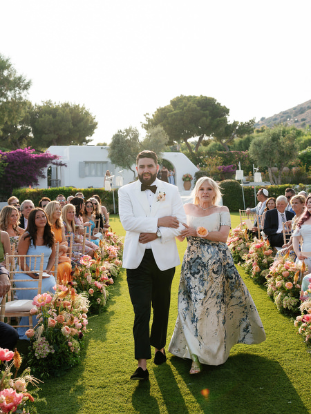 Groom and mother walking arm-in-arm down coral and blush floral-lined aisle at Island Residence