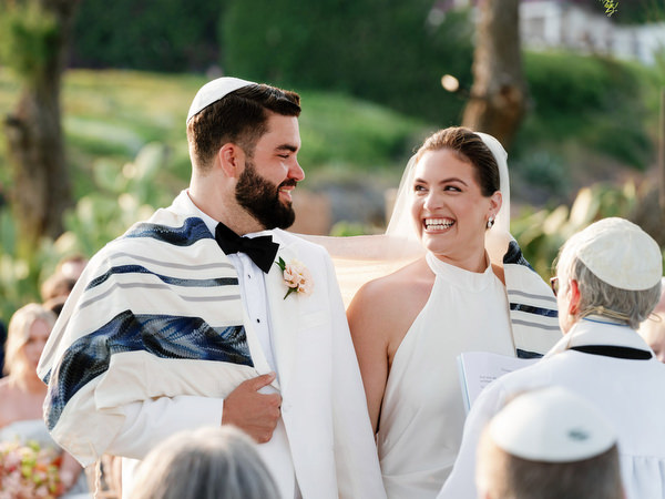 Groom wrapped in tallit smiling at bride during ceremony blessing at Island Residence