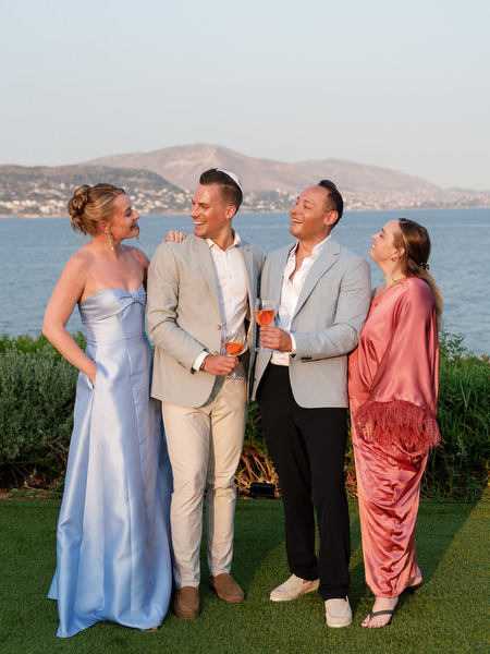 Guests in pastel formalwear holding Aperol spritz cocktails against the sea
