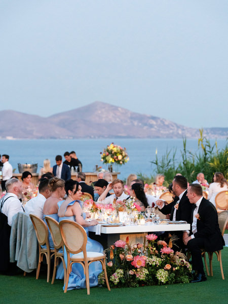 Guests seated at long banquet tables surrounded by coral and blush florals with the sea beyond