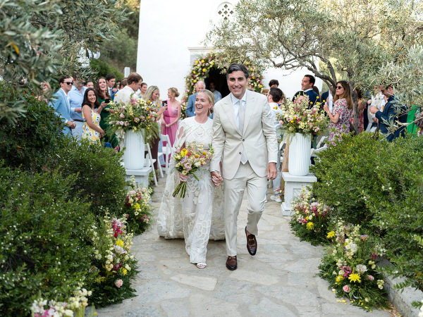 Bride and groom walking down olive-lined aisle after Orthodox wedding at Petra Farm Paros.