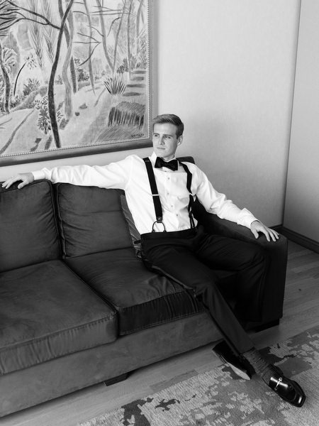 Groom seated in suspenders during wedding preparation in Athens before Catholic ceremony.