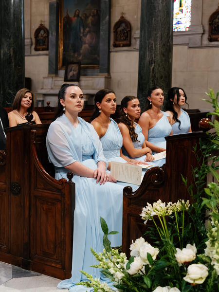 Bridesmaids in soft blue dresses seated inside Athens Catholic church during wedding ceremony.