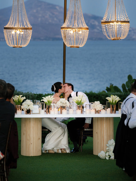 Bride and groom kissing at dinner table overlooking the Athens Riviera at night.