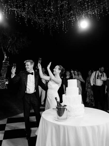 Bride and groom toasting beside wedding cake under fairy lights at Athens Riviera reception.