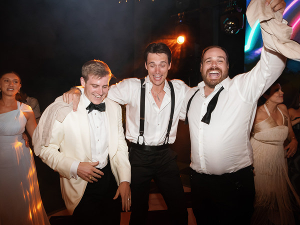 Groom and friends celebrating with arms around each other on dance floor at Athens Riviera wedding reception.