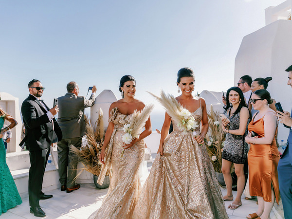Bridesmaids in gold gowns entering Santorini ceremony.