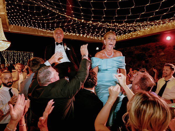 Brides mother and father lifted on chair during Jewish hora Santorini wedding.