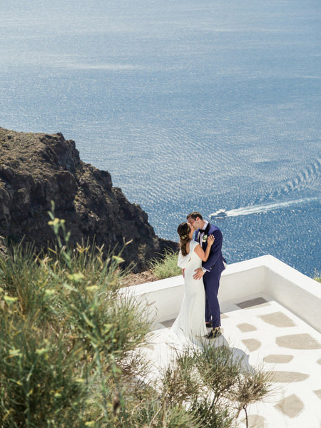 Bride and groom on Santorini cliff before Venetsanos Winery wedding.