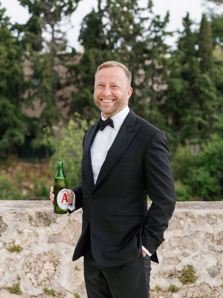 Groom in a classic black tuxedo holding a beer before his Aegina wedding