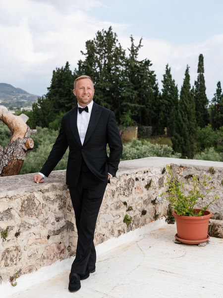 Groom posing in a black tuxedo beside a stone wall at Pyrgos Ralli in Aegina