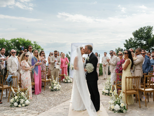 Bride and groom kissing together as guests celebrate around them in Aegina