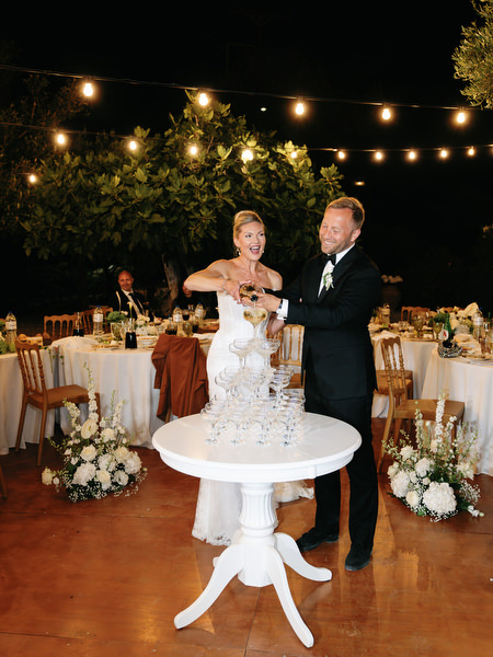 Bride and groom beside their wedding cake and champagne tower under string lights