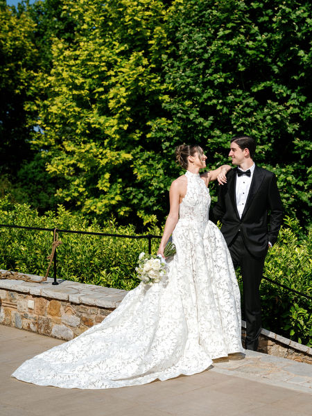 Bride and groom posing beside a stone wall with the bride holding her white bouquet