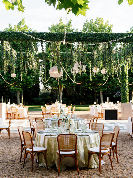 Round dinner tables arranged beneath greenery and woven lights for the outdoor reception