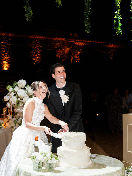 Newlyweds smiling together beside their white floral wedding cake