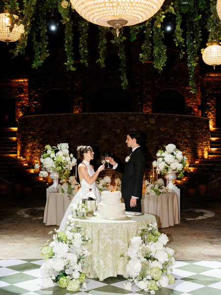 Newlyweds smiling beside their floral wedding cake during the evening reception